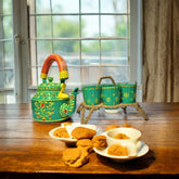 A hand-painted green aluminum kettle on a jute stand, with six green glass cups, displayed on a wooden table with cookies and a cup.